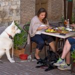 A large white and large black dog sitting next to their owners at an outdoor cafe having lunch.