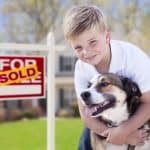 Happy Young Boy Holding Dog in front of house with for sale/sold sign