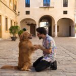 Man kneels down to his Golden Retreiver's level in a Mediterrean court yard in a respectful manner toward his dog.