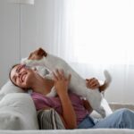 Small white terrier jumping on a young woman sitting on the couch who is his favorite person